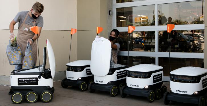 A man is putting shopping bags into a delivery robot and his colleague is...
