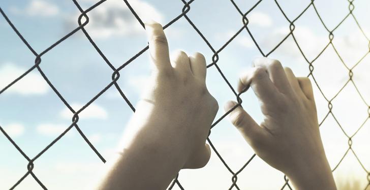 Hands holding onto a wire mesh fence, sunshine behind the fence; copyright:...