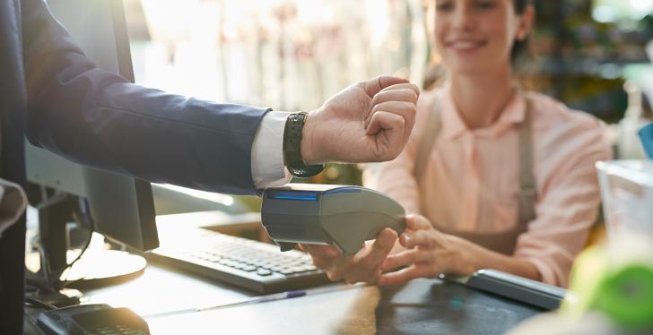 Man holds his Smartwatch to a payment device in front of smiling cashier;...