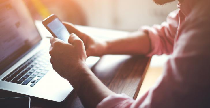 A mans hands hold a smartphone firmly in front of a laptop....