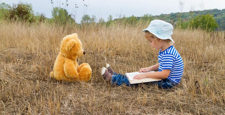 Small child sits with teddy bear and book in the grass and reads aloud...