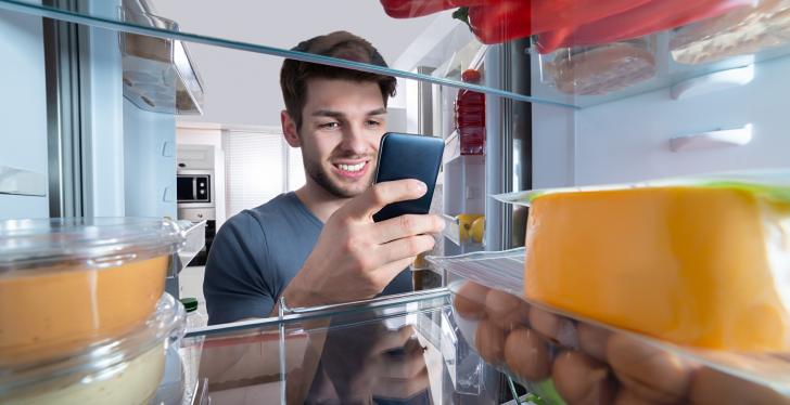 A young man looking into his fridge and holding a smartphone...