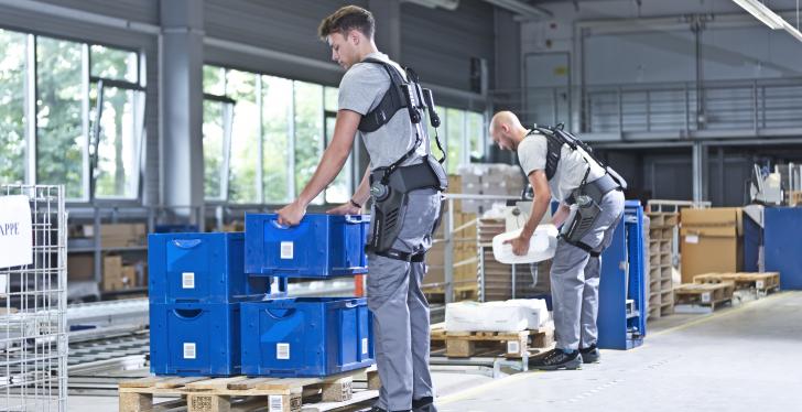 Two men lift boxes wearing exoskeletons