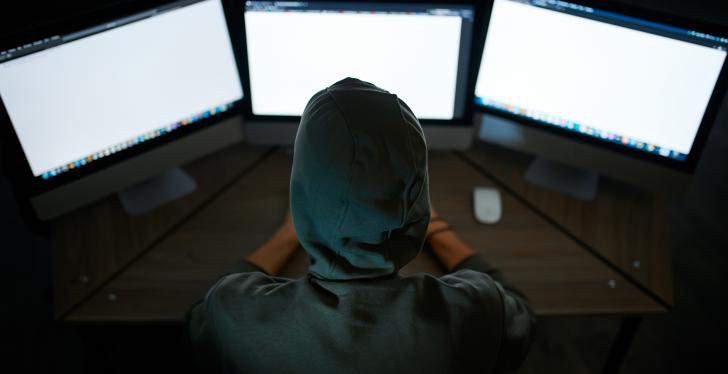 Hooded person sits in front of three computers in a dark room...
