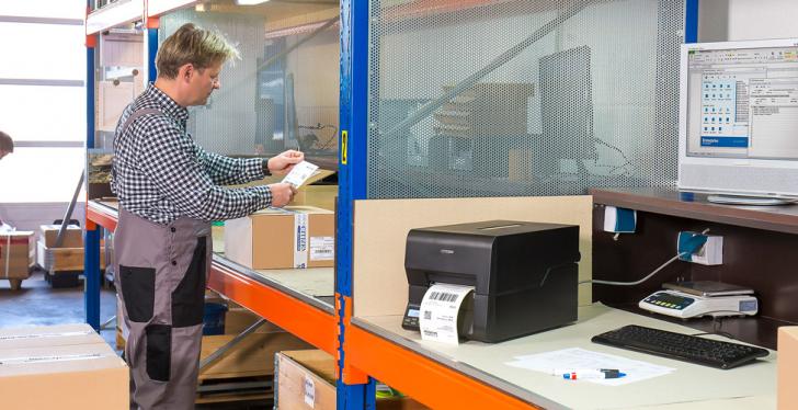 Two shipping employees stand in a hall next to a shipping label printer and...