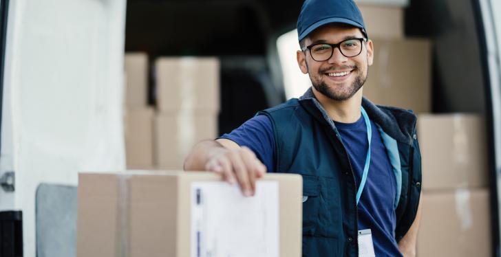 A smiling logistics worker leans against a package in front of a delivery van...