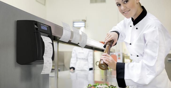 A person is seasoning a dish in a professional kitchen, with a receipt printer...