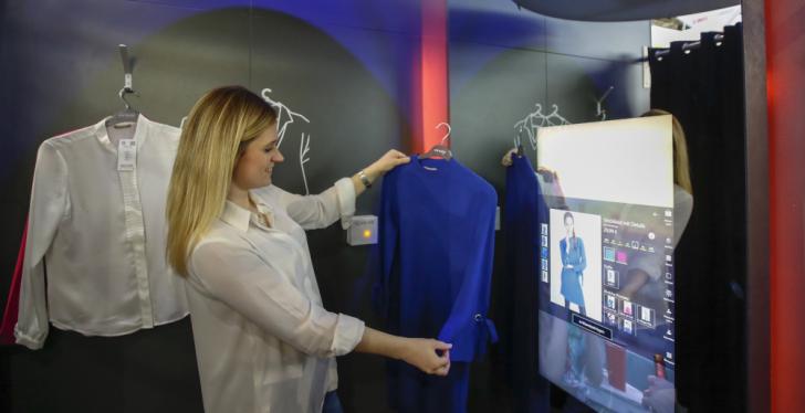 A young woman looks at a blouse in a dressing room, with an interactive digital...