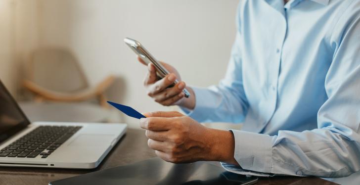 A man uses his cell phone and laptop to make a purchase. He is holding a credit...