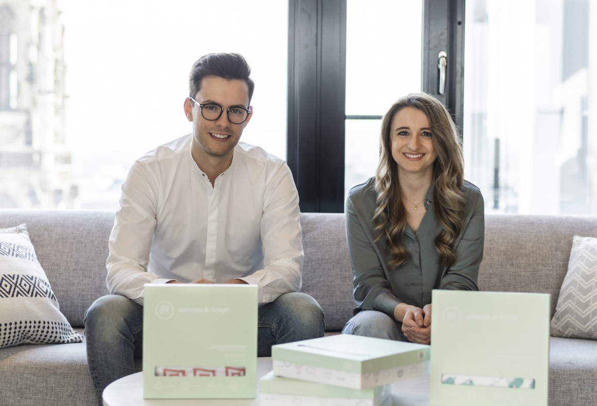 A young man and a young woman sit on a couch in front of a large window and...