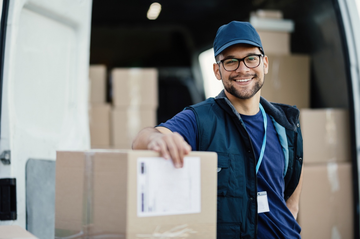 Ein Logistikmitarbeiter lehnt lächelnd an ein Paket vor einem Lieferwagen...