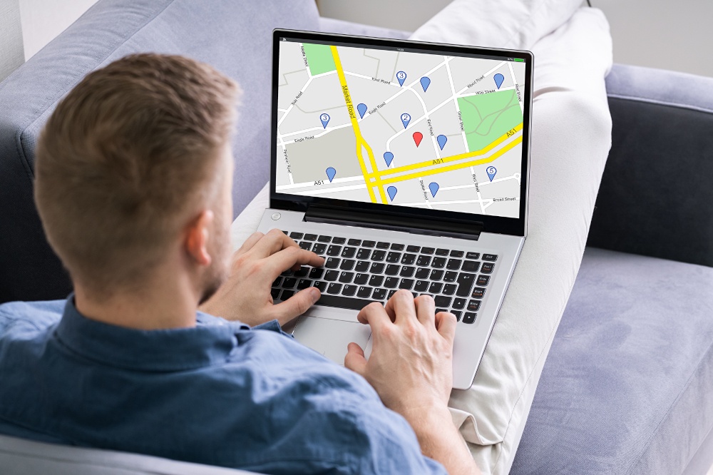 A man sitting on a couch looking at a map with locations on his laptop;...