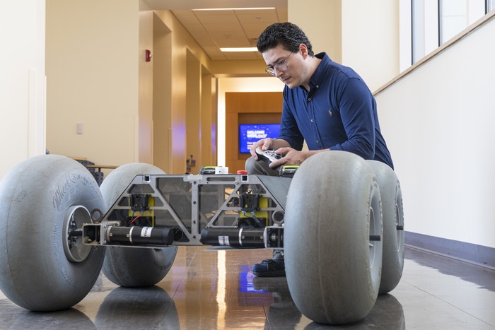 A man is kneeling in a corridor beside a robotic rover with four big wheels;...
