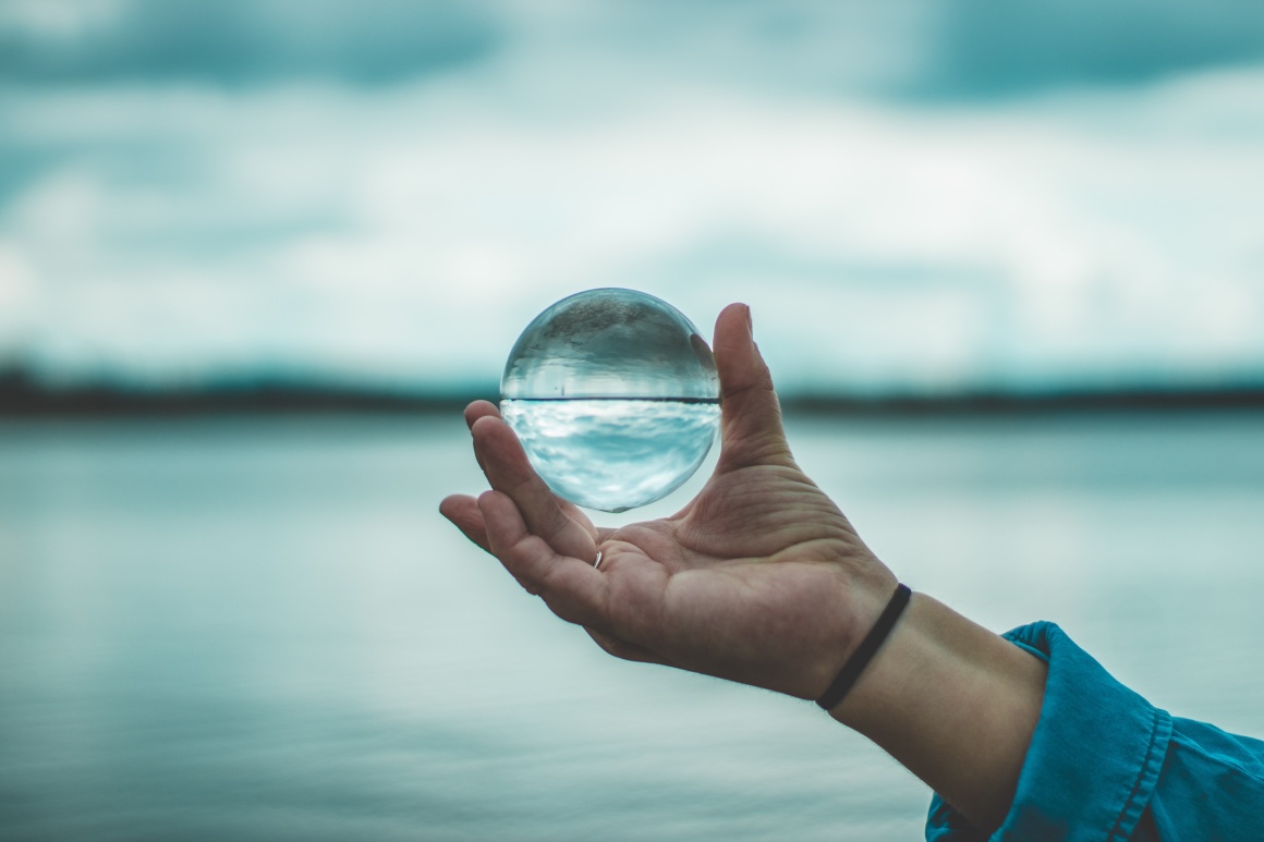 A hand holds a glass ball in which a lake and a sky are reflected...