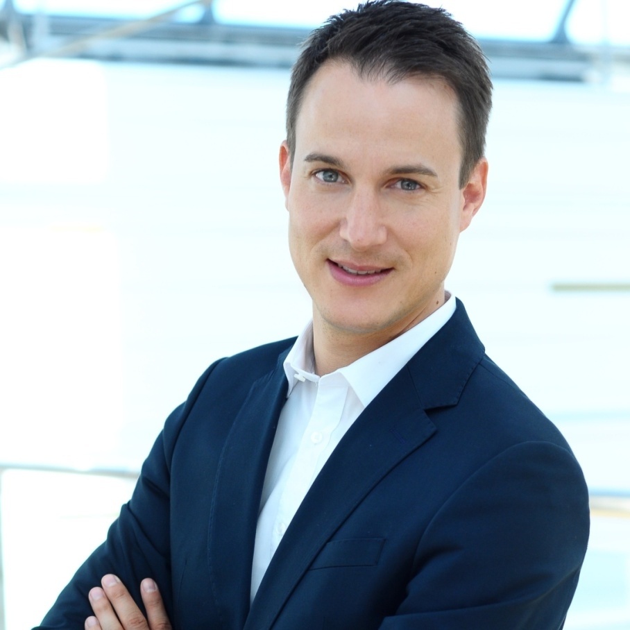 Man in suit with dark hair crosses arms and looks sideways at camera...