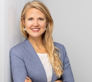 Linda Hoffmann stands with her arms folded in front of a white wall and smiles...