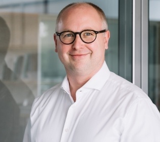 Karsten Grötecke stands in front of a glass wall and smiles into the camera....