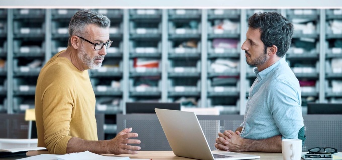 Two men are standing opposite each other and talking. One of them has a laptop...