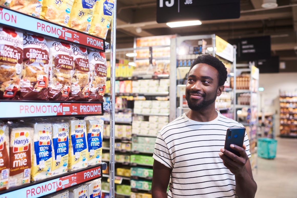 A man holding a smartphone is standing next to a supermarket shelf...