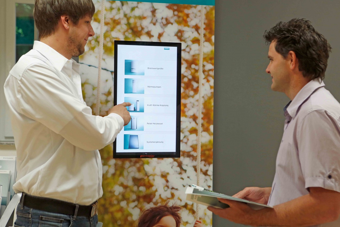 Foto: Zwei Männer vor einem digitalen Informationsbildschirm mit Produkten;...