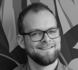 Black-and-white image of a man with glasses and beard smiling into the camera...