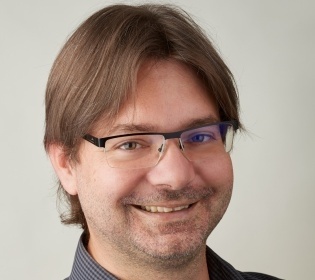 Man with glasses in grey shirt smiles into camera