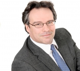 Man in suit with blue tie and glasses smiles diagonally into the camera...