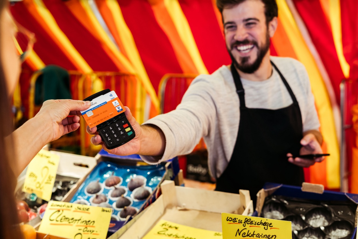 A fruit and vegetable retailer at a market stand holds out a mobile payment...