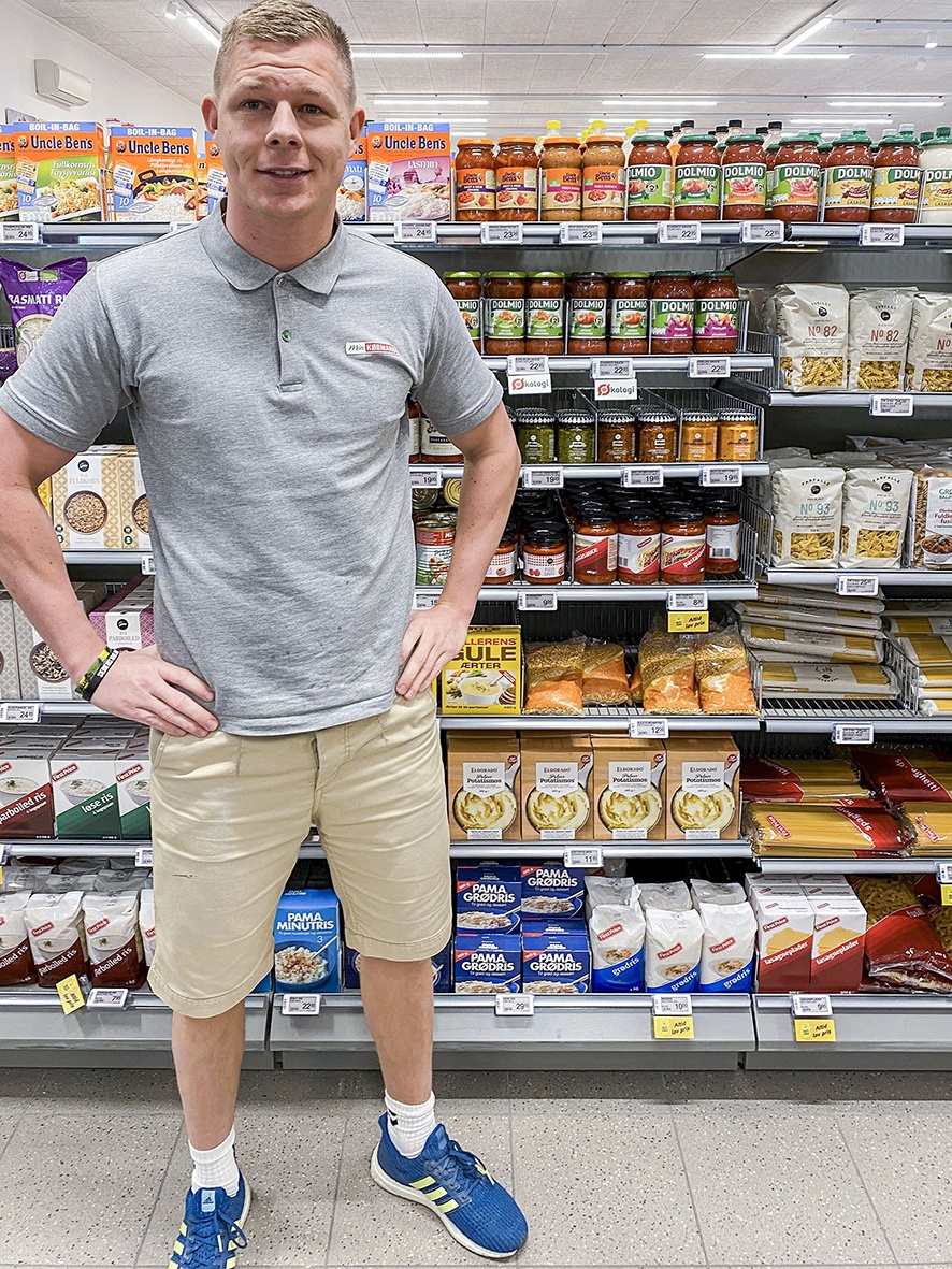 A young man standing in a small store in front of a food shelf...