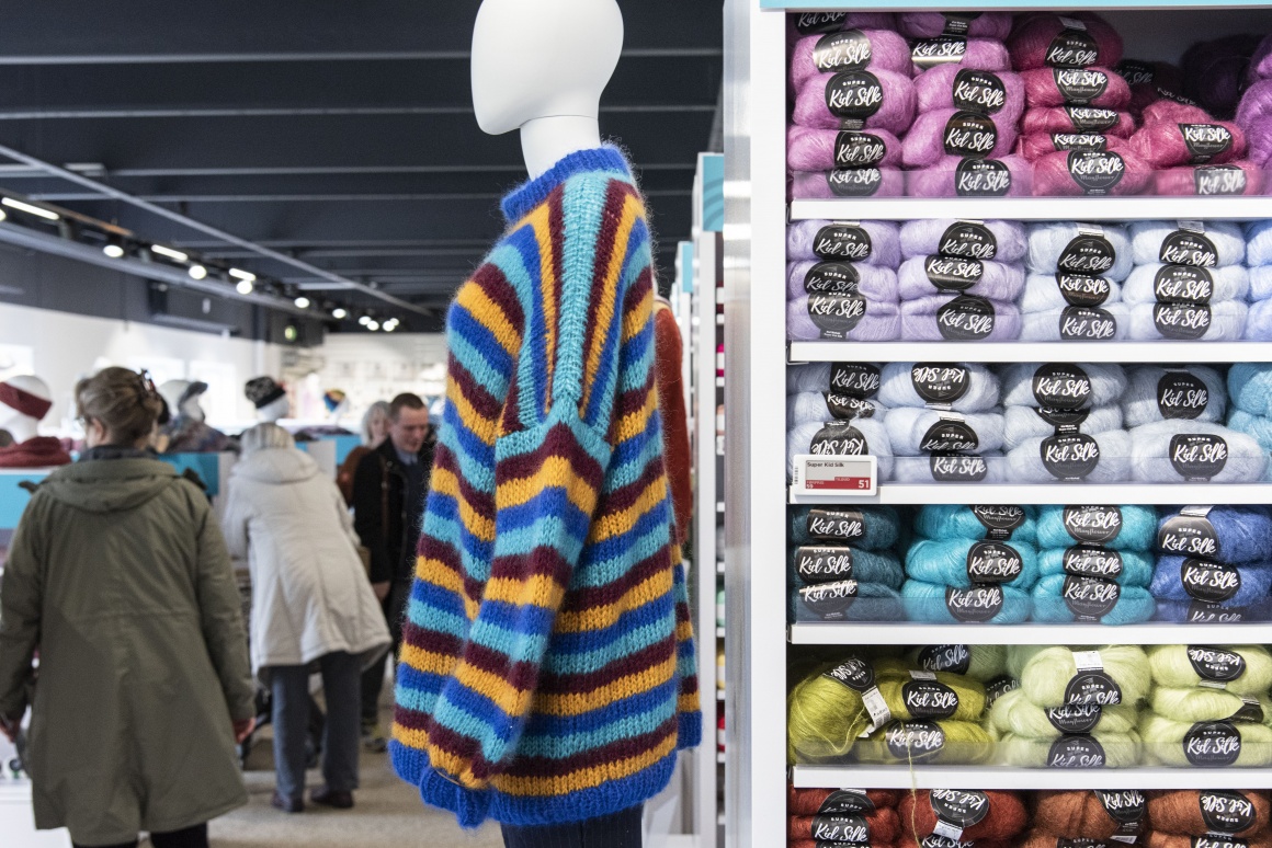 Ein Mannequin mit buntem Pullover neben einem Regal mit Wolle...