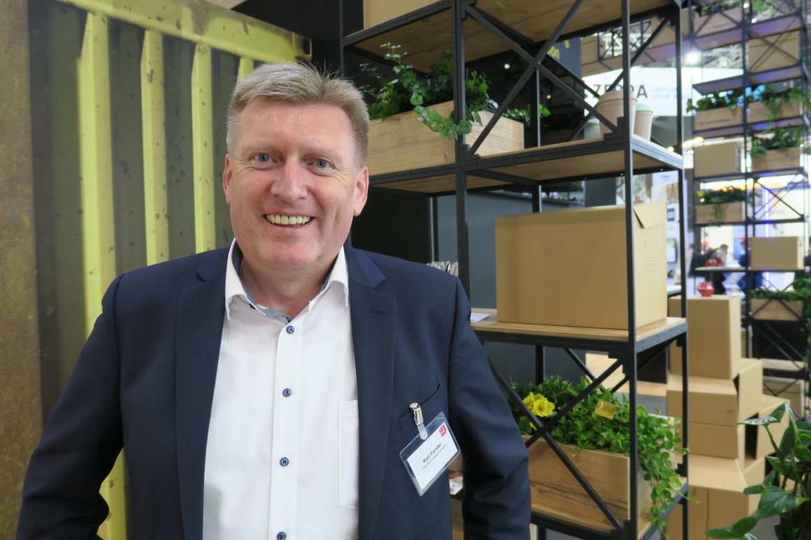 A man in a suit at an exhibition stand laughs into the camera...