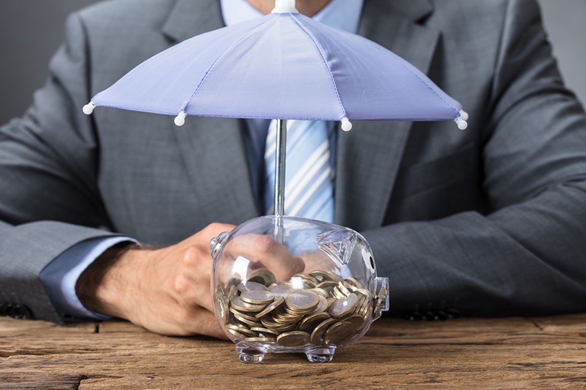 A business man sitting behind a piggybank with a small umbrella over it...