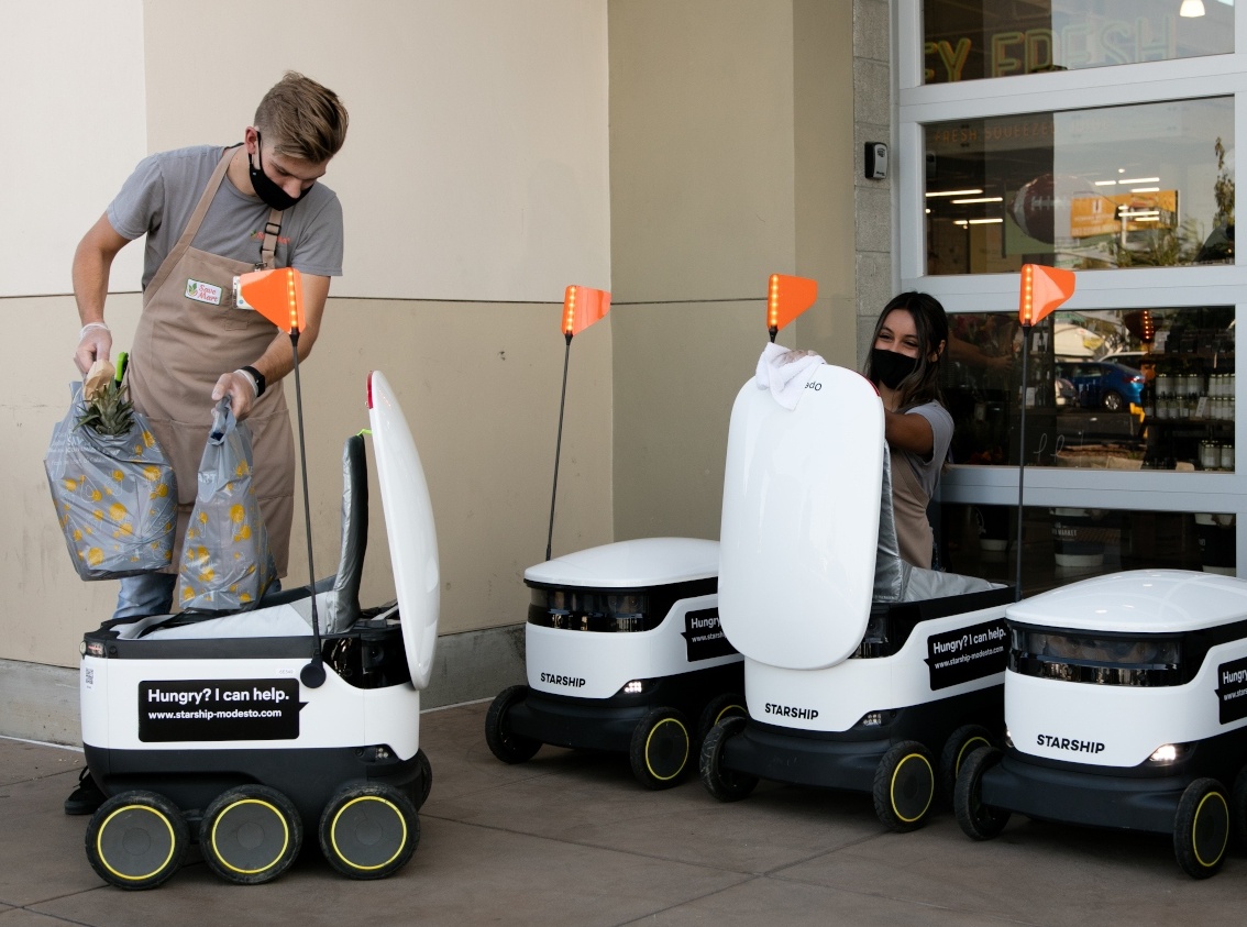 A man is putting shopping bags into a delivery robot and his colleague is...