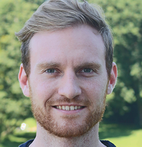 Blond man with beard smiles into the camera. In the background meadow and trees...