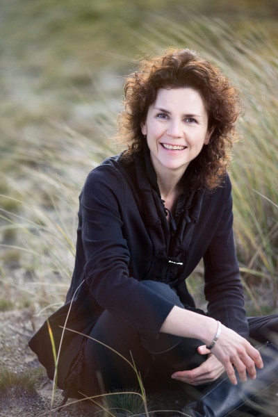 A woman with curly hair sits cross-legged on sand and smiles into the camera...