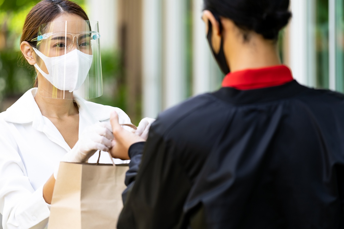 A woman with a mask is giving a brown paper bag to a person with a mask...