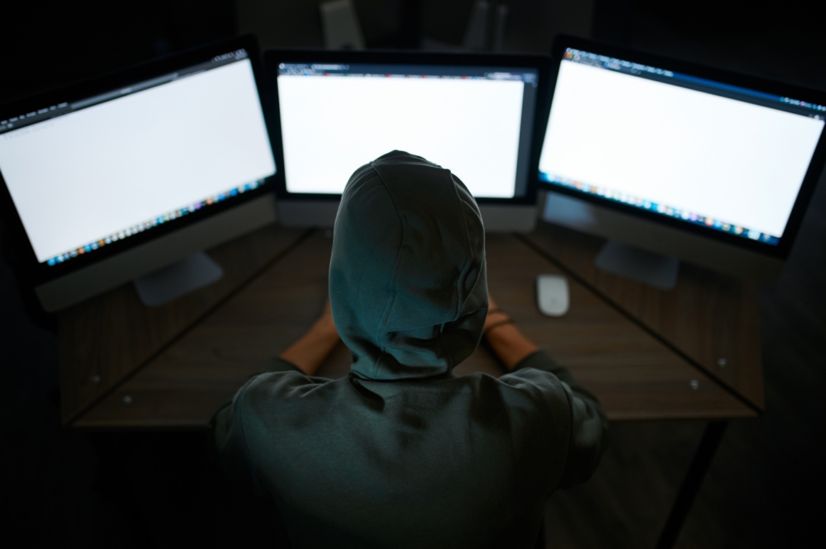 Hooded person sits in front of three computers in a dark room...