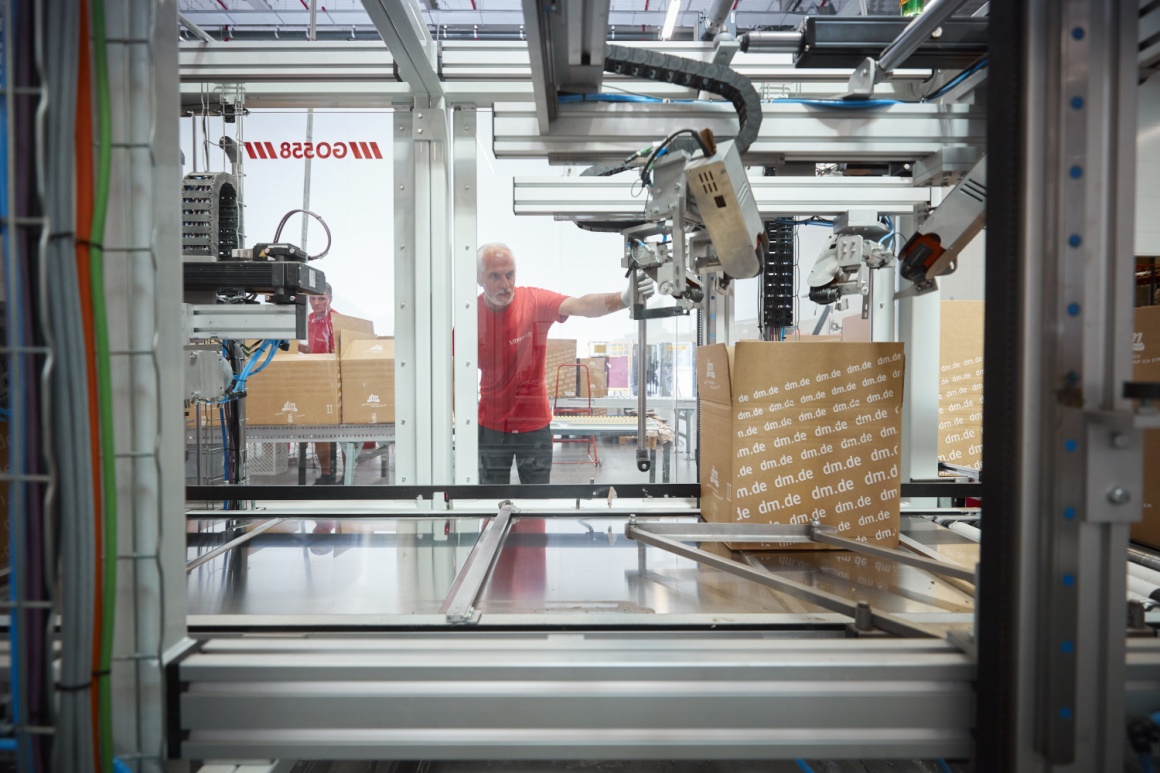 A logistics worker watches a packaging machine at work through a pane of glass;...